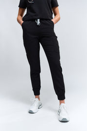 A person wearing black jogger scrub pants with multiple pockets and a drawstring waist, paired with a black scrub top and white sneakers.