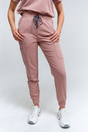 A pair of rosewood-colored jogger scrub pants with multiple pockets and a drawstring waistband, paired with white sneakers.